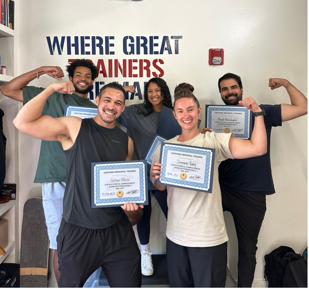 Five newly certified trainers holding their Show Up Fitness certificates in front of the Where Great Trainers Are Made wall